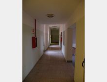 Corridoio interno lungo e stretto con pavimento in piastrelle, pareti bicolore e plafoniere a soffitto. Sulle pareti sono presenti cassette antincendio rosse ed estintori, con aperture laterali e una porta in fondo al passaggio.
