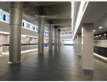 Ampio atrio interno moderno con pavimento in grandi piastrelle scure e banconi lineari lungo i lati. Colonne cilindriche scanalate sostengono un soffitto con elementi radiali decorativi, illuminate da strisce LED e finestre alte lungo il perimetro.