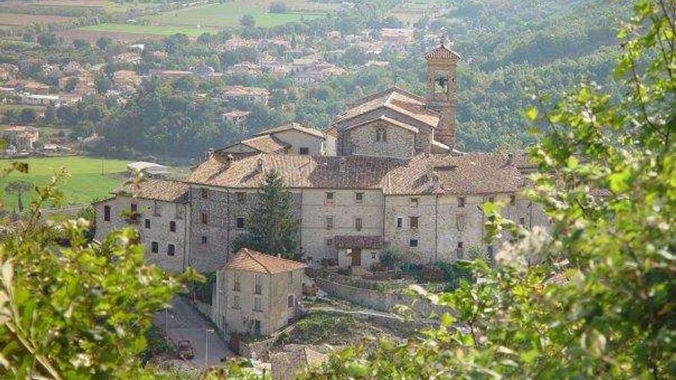 Complesso monastico in pietra con tetti in coppi e campanile, visto dall’alto tra vegetazione in primo piano. Sullo sfondo si estendono campi coltivati e un’area collinare con case sparse.