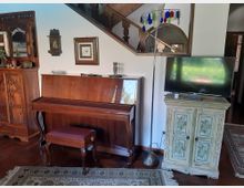 Soggiorno arredato con pianoforte verticale in legno e sgabello, accanto a un mobile dipinto con TV sopra e una lampada da terra. Sullo sfondo si vedono un grande armadio intagliato, quadri alle pareti e una scala in legno con parapetto decorato.