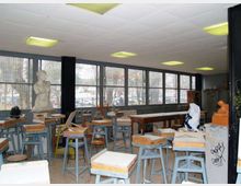 Aula-laboratorio con numerosi sgabelli e piccoli banchi da lavoro, tavoli lunghi e un busto in gesso su piedistallo. Lungo una parete ci sono grandi finestre a nastro con inferriate che affacciano sull’esterno, e un controsoffitto con plafoniere rettangolari.
