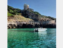 Baia costiera con acqua turchese e scogliere rocciose, con un piccolo motoscafo ancorato in primo piano. In alto sul promontorio si vede una torre circolare in pietra affiancata da un edificio basso, circondata da vegetazione mediterranea e un muro di contenimento lungo la costa.