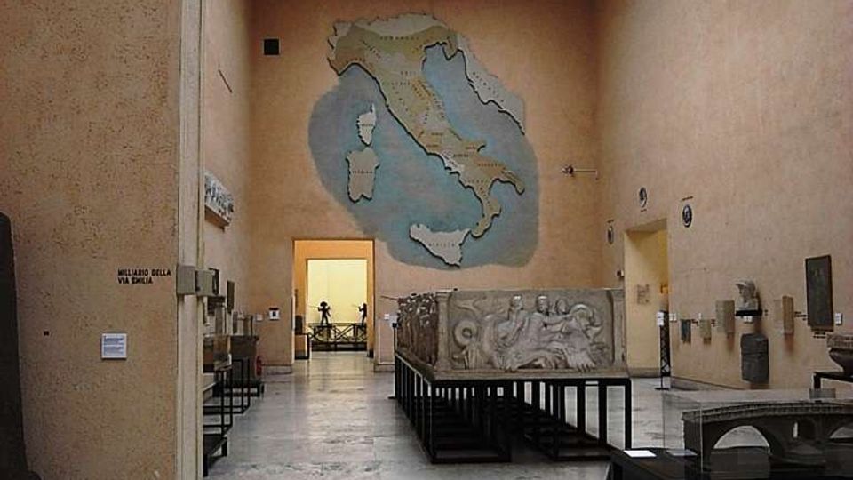 Interno di una sala museale con pareti color sabbia e lucernario a volta sul soffitto, pavimento in pietra e teche espositive con reperti lungo i lati. In fondo, una grande mappa dell’Italia dipinta sulla parete principale e un’ampia apertura che conduce a un’altra stanza.