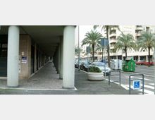 Portico urbano con lunga fila di colonne cilindriche e pavimentazione in pietra, affiancato a un marciapiede e a una strada con auto parcheggiate. Sullo sfondo si vedono palme lungo il viale, edifici residenziali alti, segnaletica stradale, un attraversamento pedonale e un parcheggio accessibile.