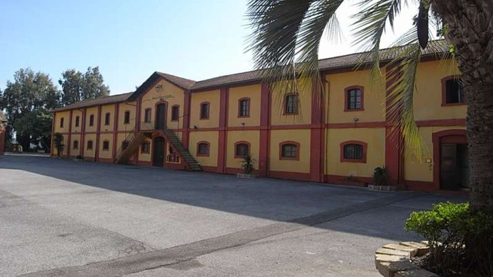 Ampio cortile asfaltato davanti a un lungo edificio a due piani in stile industriale, con facciata gialla e cornici rosse, finestre ad arco e scale esterne. In primo piano si vede una palma e ai lati altre aree verdi con alberi.
