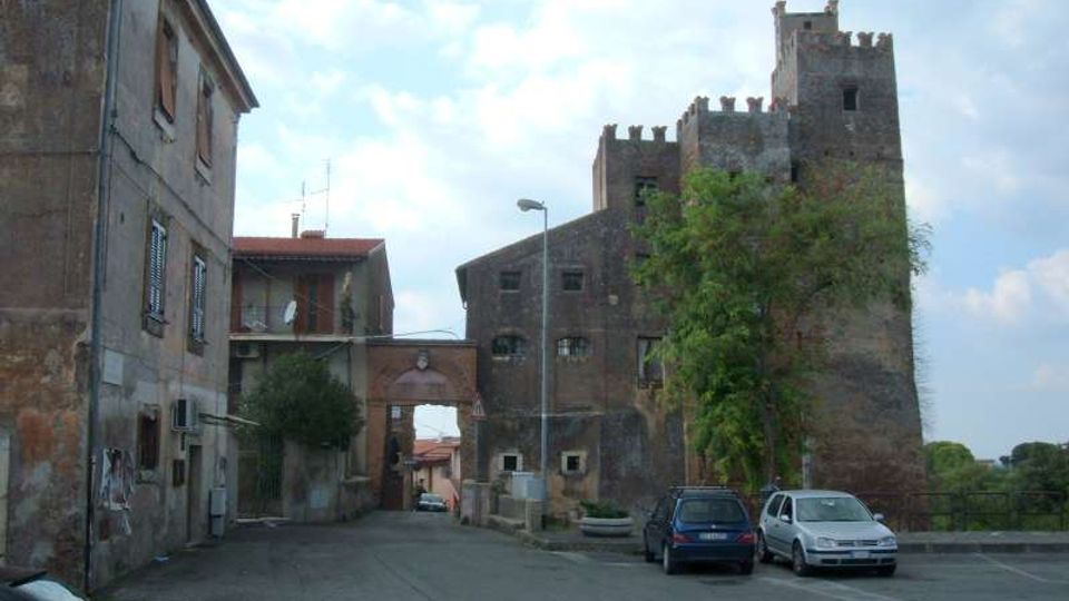 Strada di un borgo con edifici in muratura e un arco di passaggio, affiancata da un grande edificio fortificato in mattoni con torri merlate. Sul lato destro si vedono alberi e alcune auto parcheggiate lungo il bordo della carreggiata.
