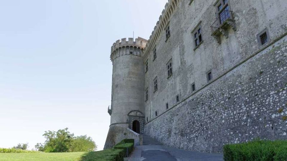 Veduta esterna di un castello in pietra con mura alte e una torre cilindrica merlata, con piccole finestre e un balconcino. Un vialetto in salita costeggiato da siepi conduce a un portale d’ingresso, con prato e alberi sul lato sinistro.