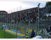 Tribuna gremita di uno stadio di calcio, con gradinate metalliche alte e recinzioni in rete e ringhiere davanti al campo. In primo piano si vedono il bordo del terreno di gioco con cartelloni pubblicitari e strutture di sicurezza lungo la curva.