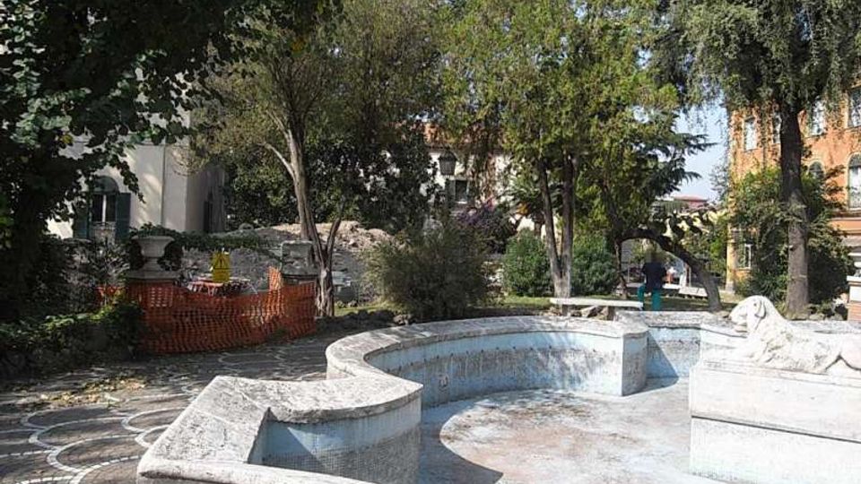 Piccolo giardino urbano con una vasca-fontana in pietra vuota dalle forme curve e basse, affiancata da un pavimento a mosaico. Alberi e cespugli circondano l’area, con edifici sullo sfondo e una recinzione arancione da cantiere su un lato.