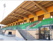 Tribuna coperta di uno stadio con struttura in cemento e travi a vista, file di sedili verdi disposte su gradinate. Scale e corrimano metallici conducono ai vari settori, con pareti giallo-arancio e ingresso vetrato alla base.