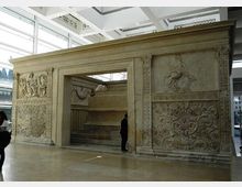 Struttura monumentale in pietra scolpita all’interno di un museo, con grande apertura centrale e pannelli decorati a bassorilievo con motivi figurativi e floreali. Ambiente moderno con pavimento lucido, ampie vetrate e soffitto a griglia metallica.