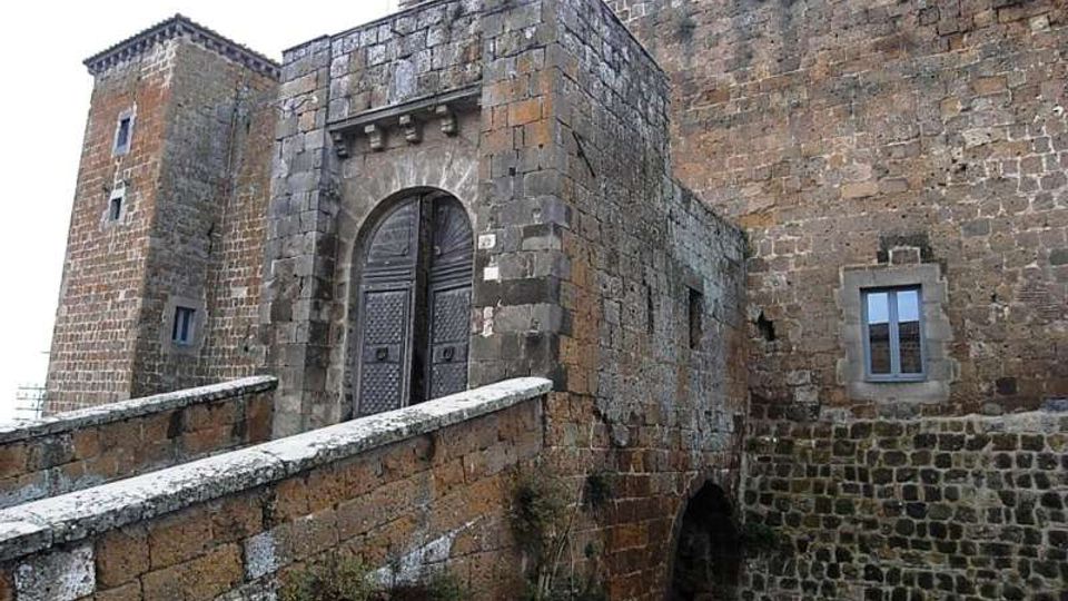 Ingresso di un castello in pietra con portale ad arco e grande porta in legno, raggiunto da un ponte in muratura. Le mura esterne massicce mostrano una torre laterale e una piccola finestra incorniciata in pietra.