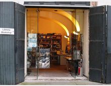Ingresso di una libreria con grandi porte scure aperte e vetrate scorrevoli, che conduce a un interno con soffitto a volte illuminato e scaffali pieni di libri. A sinistra è visibile un cartello “Libreria/Bookshop” che indica l’entrata.