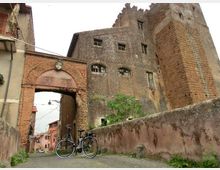 Antico complesso in mattoni con grande arco d’ingresso e torre merlata, affiancato da edifici in muratura con piccole finestre. In primo piano si vede un vicolo lastricato con un basso muro in pietra e due biciclette appoggiate.