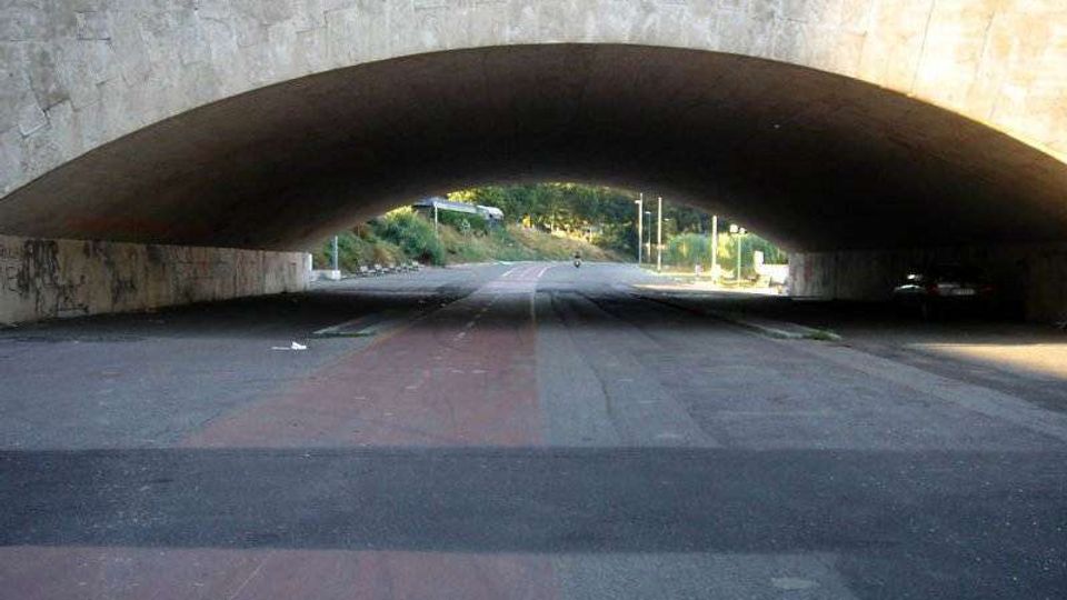 Sottopasso stradale con grande arco in pietra sopra una carreggiata ampia, con segnaletica e corsie visibili. In fondo si intravedono una strada in salita, lampioni e vegetazione ai lati.