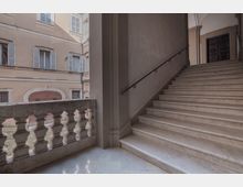 Scalinata interna in pietra chiara con corrimano in ferro che sale verso un portone scuro sotto archi, affacciata su un cortile tramite una balaustra in muratura. Sullo sfondo si vedono facciate di edificio con finestre e persiane azzurre.