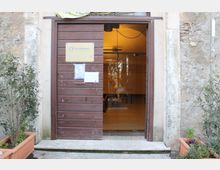 Ingresso di un edificio in pietra con grande porta in legno scorrevole e apertura che mostra un interno illuminato con pavimento piastrellato e pareti chiare. Ai lati dell’entrata ci sono vasi rettangolari con piante e una targa informativa affissa sulla porta.