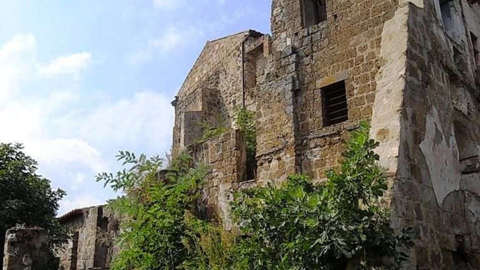 Edificio in pietra in rovina con torre a più piani, finestre vuote e intonaco scrostato. In primo piano un muretto di pietra e vegetazione spontanea che cresce lungo i resti della struttura, sotto un cielo parzialmente nuvoloso.