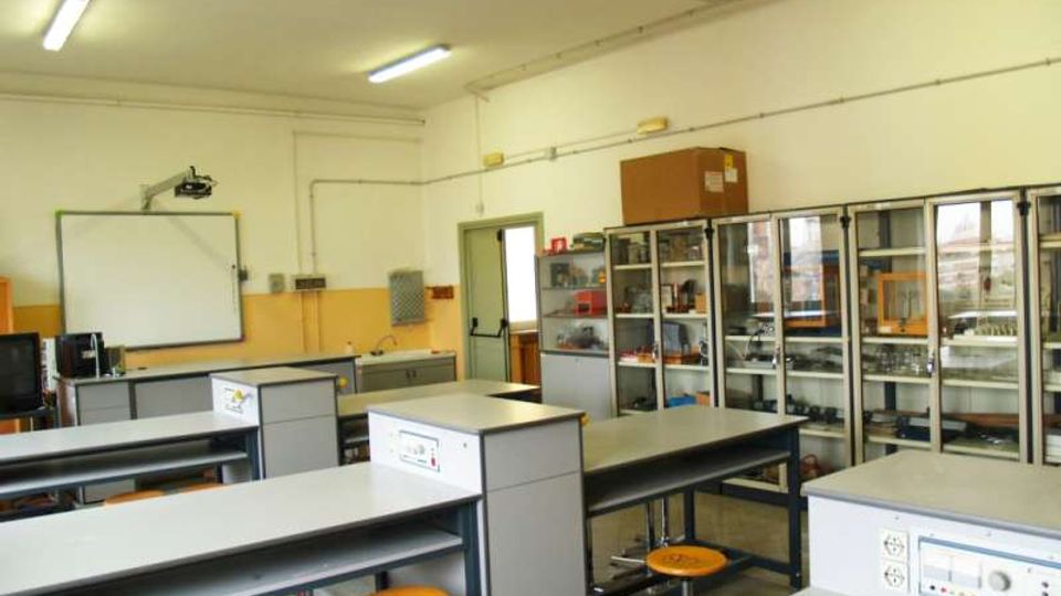 Aula laboratorio scolastico con banchi da lavoro e sgabelli gialli, illuminazione a neon e una lavagna bianca con proiettore a parete. Sul lato destro ci sono armadi vetrati con attrezzature e strumenti, e un banco con lavello sul fondo.