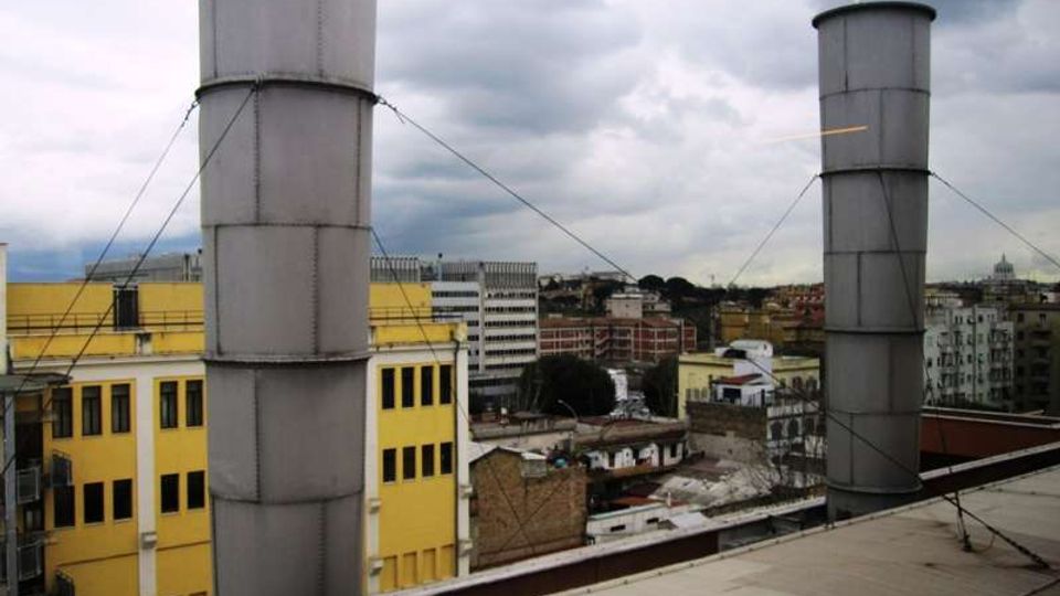 Tetto di un edificio con due alte canne fumarie cilindriche in metallo, sostenute da tiranti, affacciato su una zona urbana con palazzi e magazzini. Sullo sfondo si vedono edifici multipiano e un cielo nuvoloso.