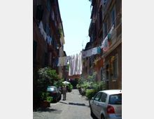 Vicolo urbano stretto con pavimentazione in sampietrini, fiancheggiato da palazzi alti con persiane e balconi. Fili per stendere il bucato attraversano la strada tra gli edifici, con auto parcheggiate e piante in vaso lungo i lati.