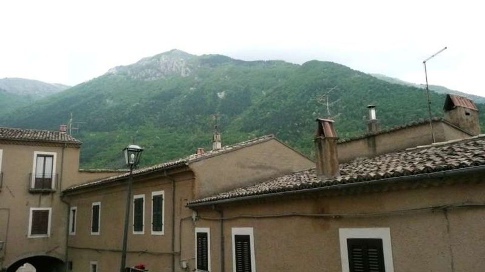 Case in muratura con facciate color ocra e tetti in coppi, con finestre a persiane scure e un piccolo passaggio ad arco. Sullo sfondo si vedono montagne verdi boscose sotto un cielo chiaro.