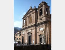 Facciata di una grande chiesa in mattoni con portali scuri, finestre ad arco e campanile laterale a loggia, vista da una piazza. In primo piano si vede una statua su piedistallo, con edifici residenziali e colline sullo sfondo.