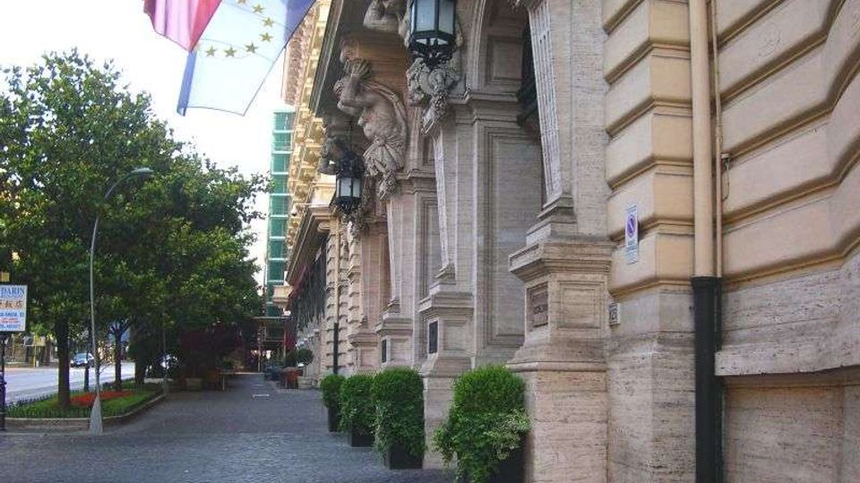 Facciata di un edificio storico in pietra con colonne e mensole scolpite, lanterne appese e bandiere esposte sopra un ampio marciapiede lastricato. Lungo la strada alberata si vedono grandi fioriere con cespugli e la carreggiata sul lato sinistro.