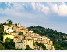 Borgo collinare con case in pietra e facciate color pastello disposte a terrazze, con tetti in coppi e un campanile che spicca tra gli edifici. Intorno si vedono pendii verdi alberati e un ampio cielo parzialmente nuvoloso.