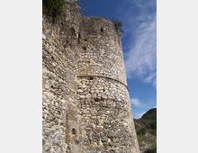 Torre cilindrica in pietra di una fortificazione, vista dal basso, con muratura irregolare e piccole aperture lungo la sommità. La struttura sorge su un pendio roccioso con vegetazione rada, sotto un cielo azzurro con nuvole sottili.