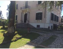 Villa su due piani con facciata chiara, finestre con persiane scure e ingresso ad arco con colonne, raggiunto da una breve scalinata. Davanti si vede un giardino con prato, palme e un vialetto carrabile in pietra che conduce all’edificio.