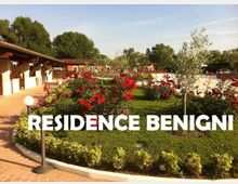 Giardino curato di una residence con prato verde, aiuole di rose rosse e piccoli alberi, circondato da vialetti in mattoni con lampioni sferici. Sullo sfondo si vedono edifici bassi con portici e tetti in tegole rosse, e una zona piscina recintata.