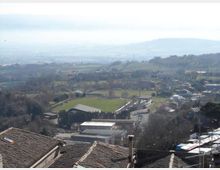 Vista panoramica dall’alto su una valle collinare con campi e piccoli insediamenti; al centro si vede uno stadio o campo sportivo ovale con tribune e impianti. In primo piano compaiono tetti in tegole, mentre sullo sfondo si estendono colline e montagne in lontananza.