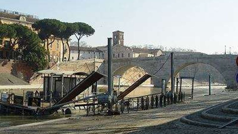 Veduta di un ponte storico in pietra ad arcate su un fiume, con una banchina pavimentata in primo piano e una struttura metallica con rampe d’accesso lungo la riva. Sullo sfondo si vedono edifici e alberi sul lungofiume.