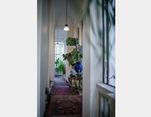 Corridoio interno luminoso con pareti bianche e soffitto alto a volta, illuminato da lampade a sospensione. Sul pavimento ci sono tappeti persiani e, lungo il lato, un tavolino in legno con vasi e grandi piante vicino a finestre con inferriate decorative.