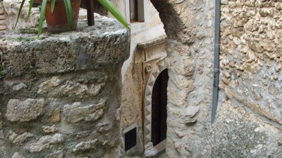 Vicolo in pietra in un borgo storico, con una scalinata acciottolata che scende sotto un arco in muratura collegato a una parete in pietra grezza. Si vede una piccola porta ad arco con cornice decorata e un muretto in pietra con vaso di piante.