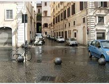 Strada urbana stretta con pavimentazione in pietra bagnata, fiancheggiata da edifici storici in muratura con finestre e archi al piano terra. In primo piano si vedono dissuasori sferici, una bicicletta e veicoli parcheggiati lungo i lati della via.