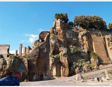 Rupe rocciosa con pareti di tufo erose e resti di mura e strutture in pietra sulla sommità, con alberi e vegetazione che crescono tra le fessure. In basso si vede una strada ai piedi della roccia e un cielo sereno sullo sfondo.