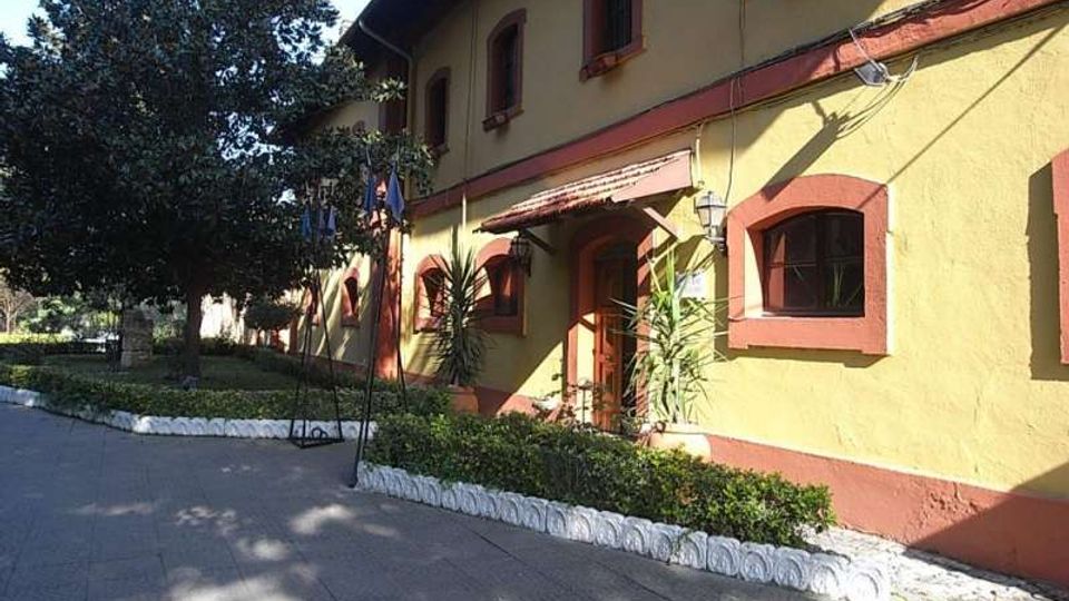 Edificio a due piani con facciata giallo ocra e cornici rosse intorno a finestre e porte, con piccolo portico d’ingresso. Davanti c’è un vialetto pavimentato con aiuole bordate di bianco, bandiere su aste e alberi che proiettano ombre sul suolo.