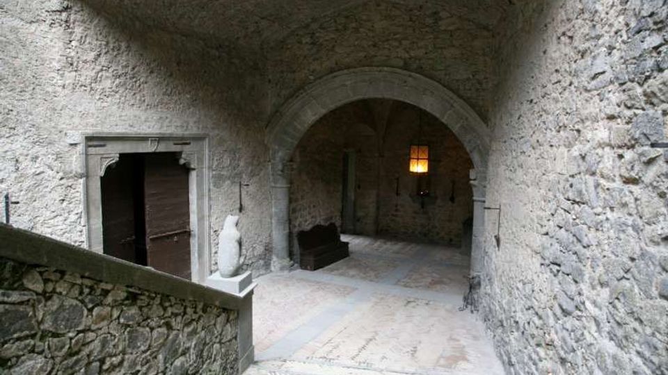 Cortile interno in pietra con soffitto a volta e grande arco che conduce a un corridoio coperto, illuminato da una piccola finestra. Sulla sinistra si vede una porta in legno incorniciata da pietra e una breve scalinata con muretto in muratura.
