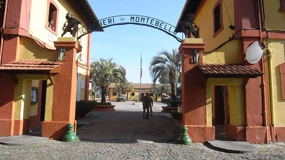 Ingresso monumentale a un complesso militare, con due edifici simmetrici giallo e rosso e un arco con la scritta “... di Montebello” tra due pilastri decorati. Davanti c’è una pavimentazione in ciottoli e oltre l’arco si vede un viale con palme e una bandiera al centro del cortile.