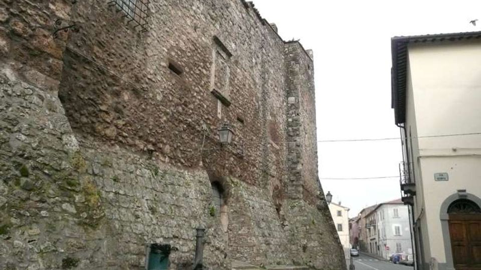 Muro esterno di una fortificazione in pietra e mattoni con piccole feritoie e finestre grate, affiancato da una strada urbana. In primo piano si vede una piccola fontana in pietra ai piedi della muratura, con lampioni e edifici moderni sul lato opposto.