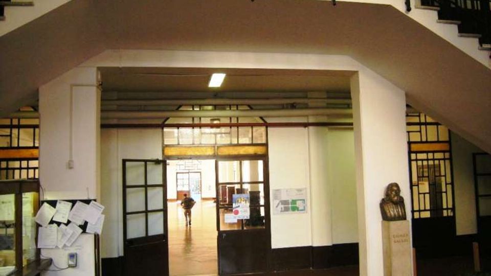 Atrio interno di un edificio pubblico con scalinate simmetriche ai lati e ringhiere nere, pareti bianche e pavimento scuro. Al centro si apre un passaggio con porte e vetrate a griglia che conduce a un corridoio illuminato; ai lati sono presenti bacheche e una statua su piedistallo.