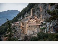 Monastero in pietra con tetti in tegole rosse costruito su una parete rocciosa a strapiombo, con torre cilindrica e arcate. Intorno si vedono scogliere e boschi di montagna, con vette sullo sfondo.