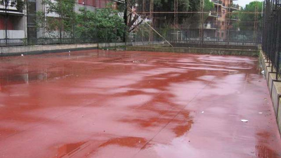 Campo sportivo all’aperto con pavimentazione rossa bagnata dalla pioggia e pozzanghere, circondato da alte recinzioni metalliche. Sullo sfondo si vedono alberi e edifici residenziali e industriali ai margini dell’area.