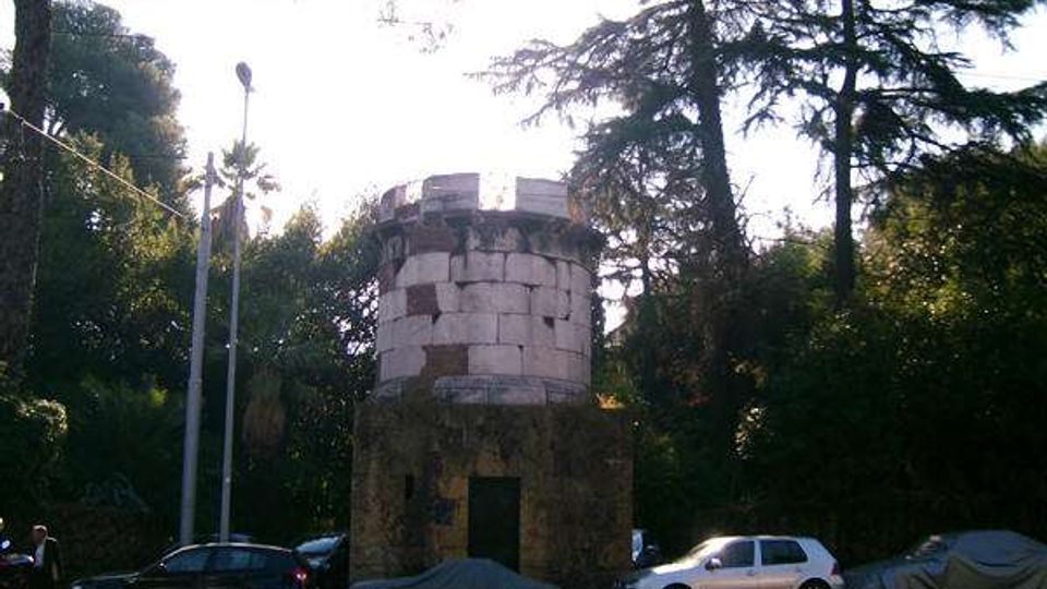 Piccola torre cilindrica in muratura con coronamento merlato, situata al margine di una strada con auto parcheggiate. Intorno ci sono alberi alti e vegetazione fitta, con lampioni stradali e luce del sole filtrata tra le chiome.