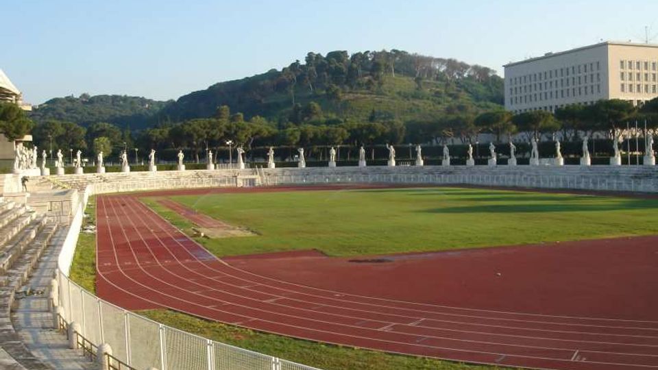 Stadio all’aperto con pista di atletica rossa che circonda un campo in erba, gradinate in pietra e recinzione metallica lungo il bordo. Sullo sfondo si vedono una fila di statue e alberi, una collina verde e un edificio rettangolare a più piani.