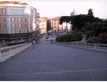 Ampia scalinata in pietra con balaustre che scende verso una piazza urbana, circondata da palazzi storici e una strada con auto. Sullo sfondo si vedono alberi di un parco e una cupola in lontananza.