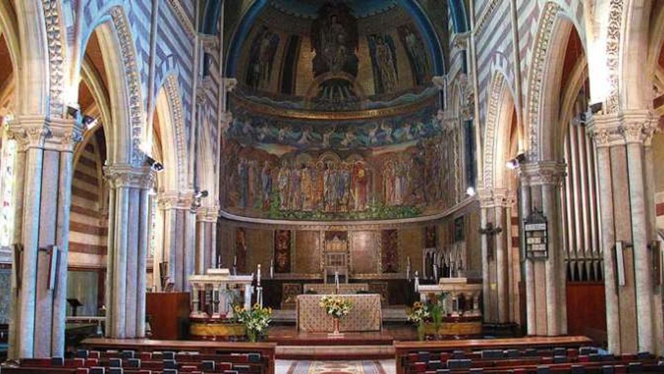 Interno di una chiesa con navata centrale e arcate a tutto sesto su colonne, pareti a fasce orizzontali e soffitto a volta. In fondo si vede il presbiterio rialzato con altare e grande abside decorata da affreschi e mosaici.