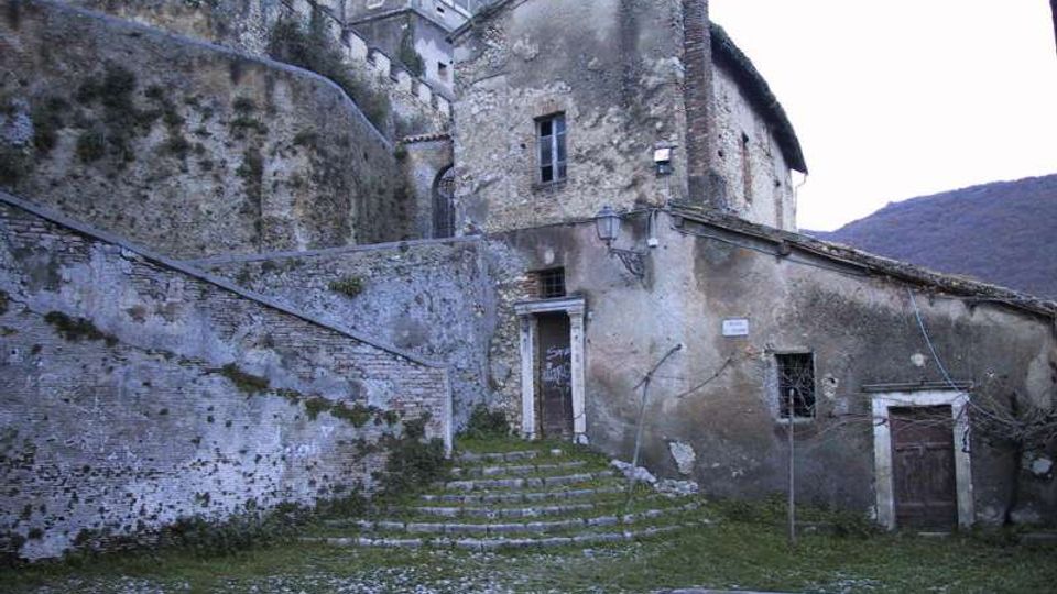 Antico complesso in pietra con edifici addossati a un pendio, mura in laterizio e una scalinata che conduce a un portale centrale. In primo piano si vede una piccola piazza erbosa con muretti e, sullo sfondo, il profilo delle colline.